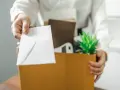 Une main tend une enveloppe blanche, comme un exemple de lettre de démission, au-dessus d'une boîte remplie d'objets personnels et d'une plante verte.
