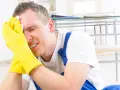 Homme en bleu de travail, gants jaunes, essuyant son front avec un mouchoir. Il semble souffrir, peut-être suite à un refus d'indemnité temporaire d'inaptitude.