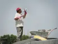 Un ouvrier boit de l'eau sous un ciel gris, trop chaud pour travailler. Il porte une casquette rouge et des gants roses.