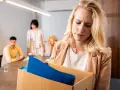 Femme blonde triste portant une boîte avec ses affaires, suite à un licenciement nul. Des collègues la regardent en arrière-plan.
