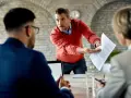 Un homme en pull rouge, visiblement en colère, tend des documents à un collègue, prêt à quitter son poste de travail après une altercation.