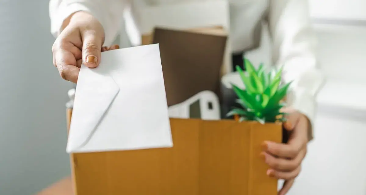 Une main tend une enveloppe blanche, comme un exemple de lettre de démission, au-dessus d'une boîte remplie d'objets personnels et d'une plante verte.