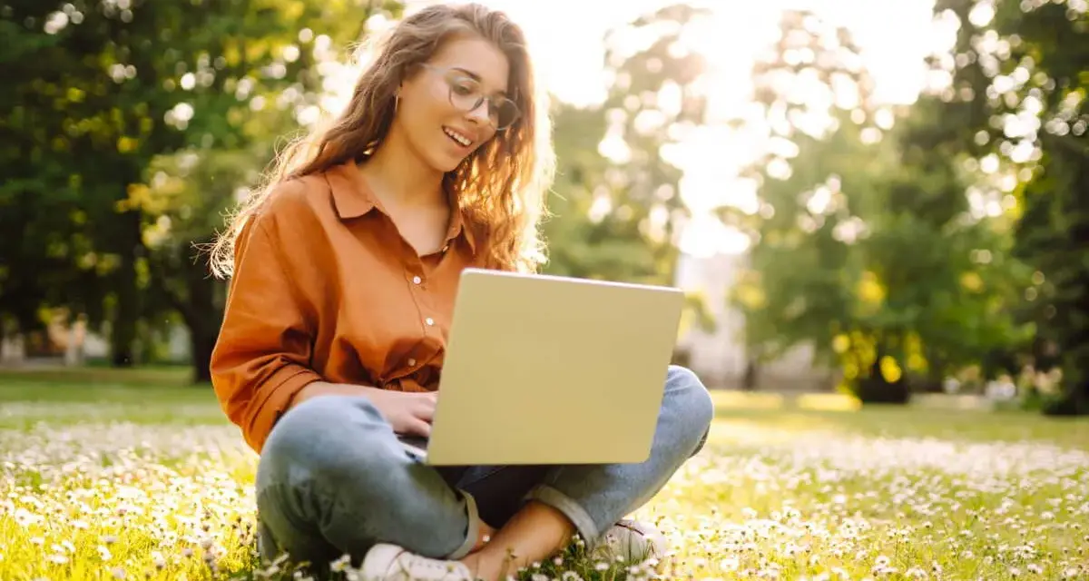 Jeune femme souriante travaillant sur son ordinateur portable dans un parc fleuri, explorant ses options pour l'âge légal pour travailler.