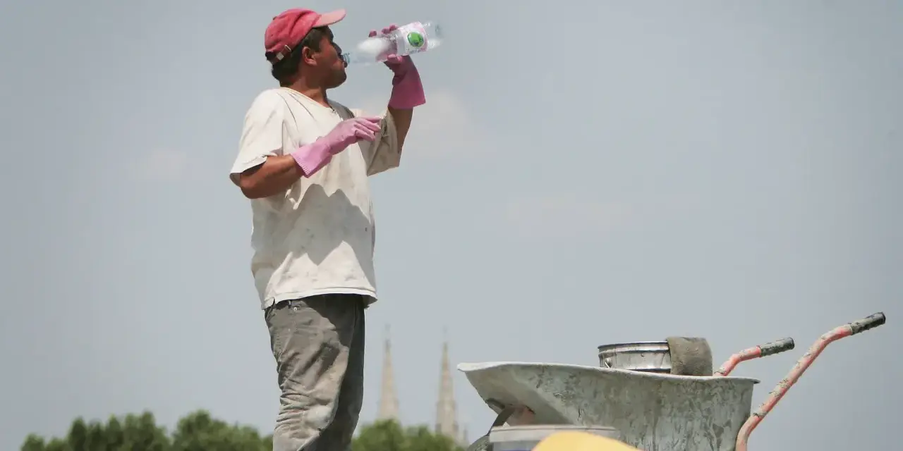 Un ouvrier boit de l'eau sous un ciel gris, trop chaud pour travailler. Il porte une casquette rouge et des gants roses.