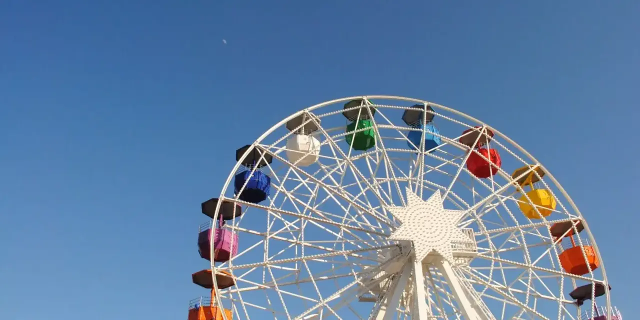 Grande roue colorée sous un ciel bleu clair, avec la lune visible. Une vue qui donne envie de monter, pour un asc cse inoubliable.