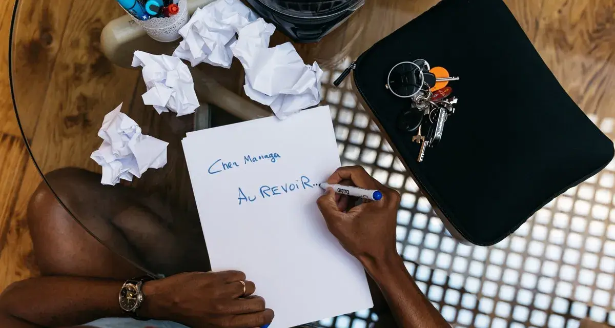 Un homme écrit "Au revoir..." sur une feuille, peut-être un mail de démission. Des brouillons froissés et des clés sont à proximité.
