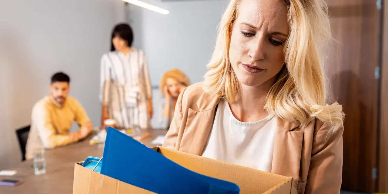 Femme blonde triste portant une boîte avec ses affaires, suite à un licenciement nul. Des collègues la regardent en arrière-plan.