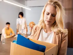 Femme blonde triste portant une boîte avec ses affaires, suite à un licenciement nul. Des collègues la regardent en arrière-plan.