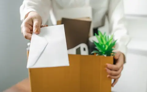 Une main tend une enveloppe blanche, comme un exemple de lettre de démission, au-dessus d'une boîte remplie d'objets personnels et d'une plante verte.