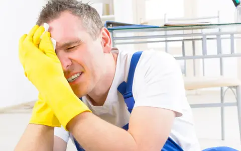 Homme en bleu de travail, gants jaunes, essuyant son front avec un mouchoir. Il semble souffrir, peut-être suite à un refus d'indemnité temporaire d'inaptitude.