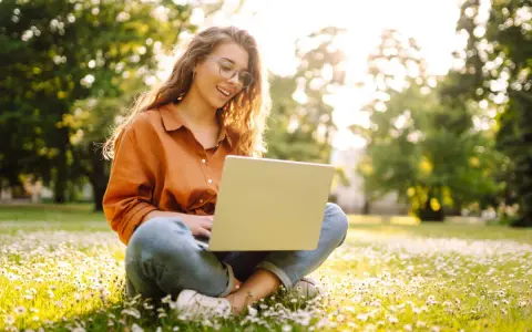 Jeune femme souriante travaillant sur son ordinateur portable dans un parc fleuri, explorant ses options pour l'âge légal pour travailler.
