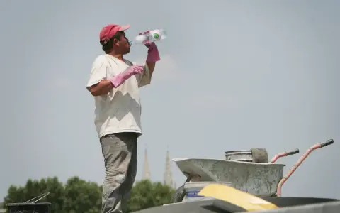 Un ouvrier boit de l'eau sous un ciel gris, trop chaud pour travailler. Il porte une casquette rouge et des gants roses.