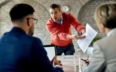 Un homme en pull rouge, visiblement en colère, tend des documents à un collègue, prêt à quitter son poste de travail après une altercation.