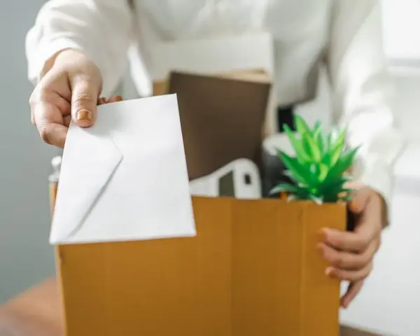 Une main tend une enveloppe blanche, comme un exemple de lettre de démission, au-dessus d'une boîte remplie d'objets personnels et d'une plante verte.