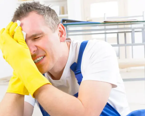 Homme en bleu de travail, gants jaunes, essuyant son front avec un mouchoir. Il semble souffrir, peut-être suite à un refus d'indemnité temporaire d'inaptitude.