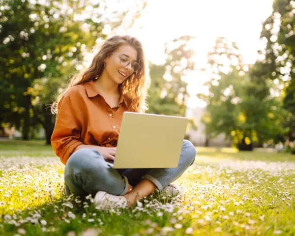 Jeune femme souriante travaillant sur son ordinateur portable dans un parc fleuri, explorant ses options pour l'âge légal pour travailler.