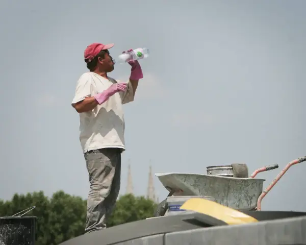 Un ouvrier boit de l'eau sous un ciel gris, trop chaud pour travailler. Il porte une casquette rouge et des gants roses.