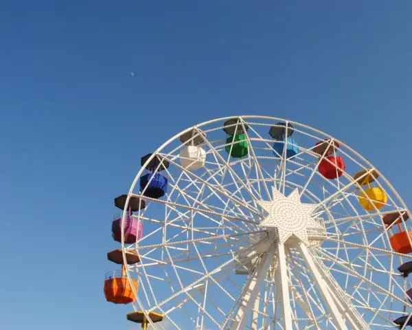 Grande roue colorée sous un ciel bleu clair, avec la lune visible. Une vue qui donne envie de monter, pour un asc cse inoubliable.