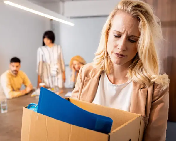 Femme blonde triste portant une boîte avec ses affaires, suite à un licenciement nul. Des collègues la regardent en arrière-plan.