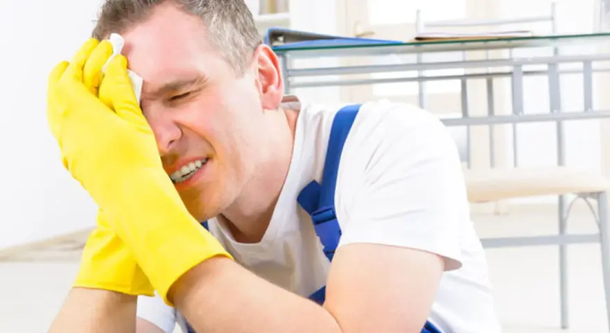 Homme en bleu de travail, gants jaunes, essuyant son front avec un mouchoir. Il semble souffrir, peut-être suite à un refus d'indemnité temporaire d'inaptitude.
