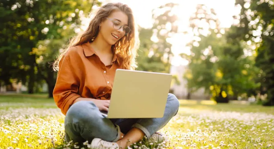 Jeune femme souriante travaillant sur son ordinateur portable dans un parc fleuri, explorant ses options pour l'âge légal pour travailler.