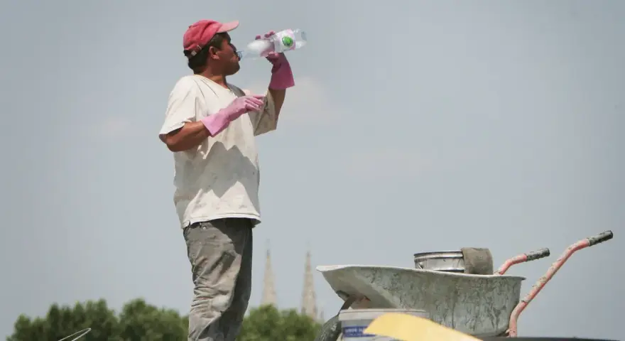 Un ouvrier boit de l'eau sous un ciel gris, trop chaud pour travailler. Il porte une casquette rouge et des gants roses.