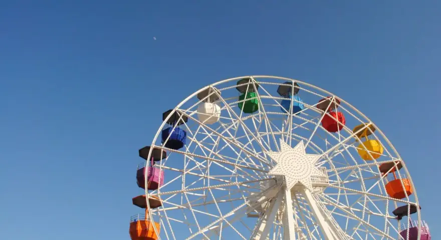 Grande roue colorée sous un ciel bleu clair, avec la lune visible. Une vue qui donne envie de monter, pour un asc cse inoubliable.