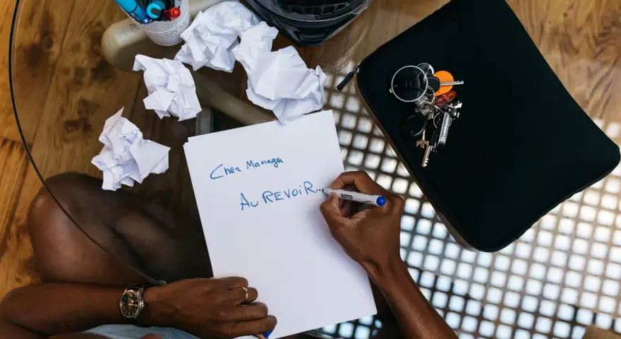 Un homme écrit "Au revoir..." sur une feuille, peut-être un mail de démission. Des brouillons froissés et des clés sont à proximité.