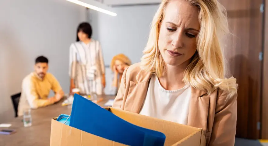 Femme blonde triste portant une boîte avec ses affaires, suite à un licenciement nul. Des collègues la regardent en arrière-plan.