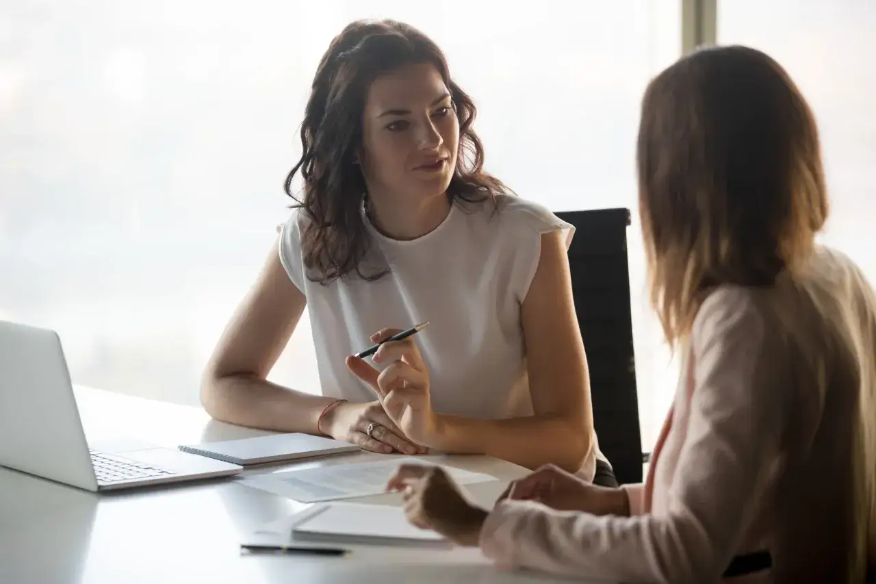 Deux femmes discutent autour d'une table, un ordinateur portable et des documents &agrave; port&eacute;e de main. Une sc&egrave;ne typique de trame entretien professionnel.