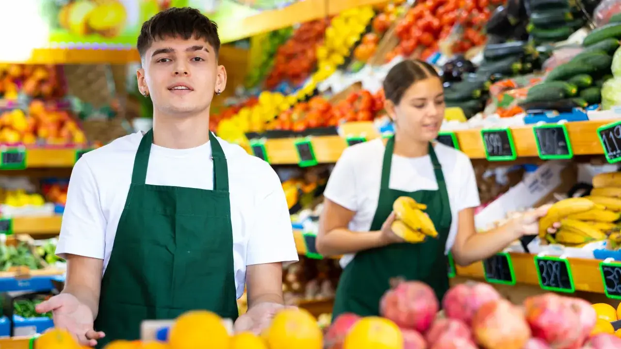 Un jeune homme de 17 ans, en tablier vert, travaille dans un rayon fruits et légumes. Il est entouré de fruits frais et colorés.