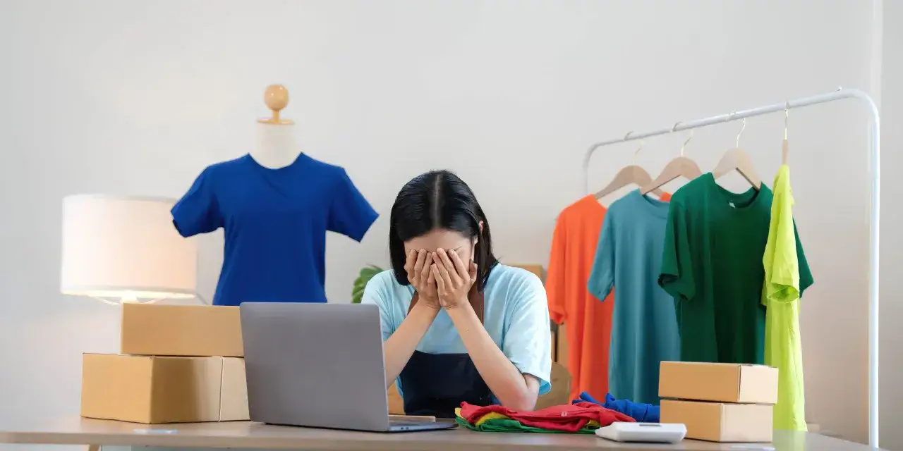 Une jeune femme désespérée devant son ordinateur, entourée de t-shirts colorés et de colis. Elle semble dépassée par la charge de travail, en sous-effectif.