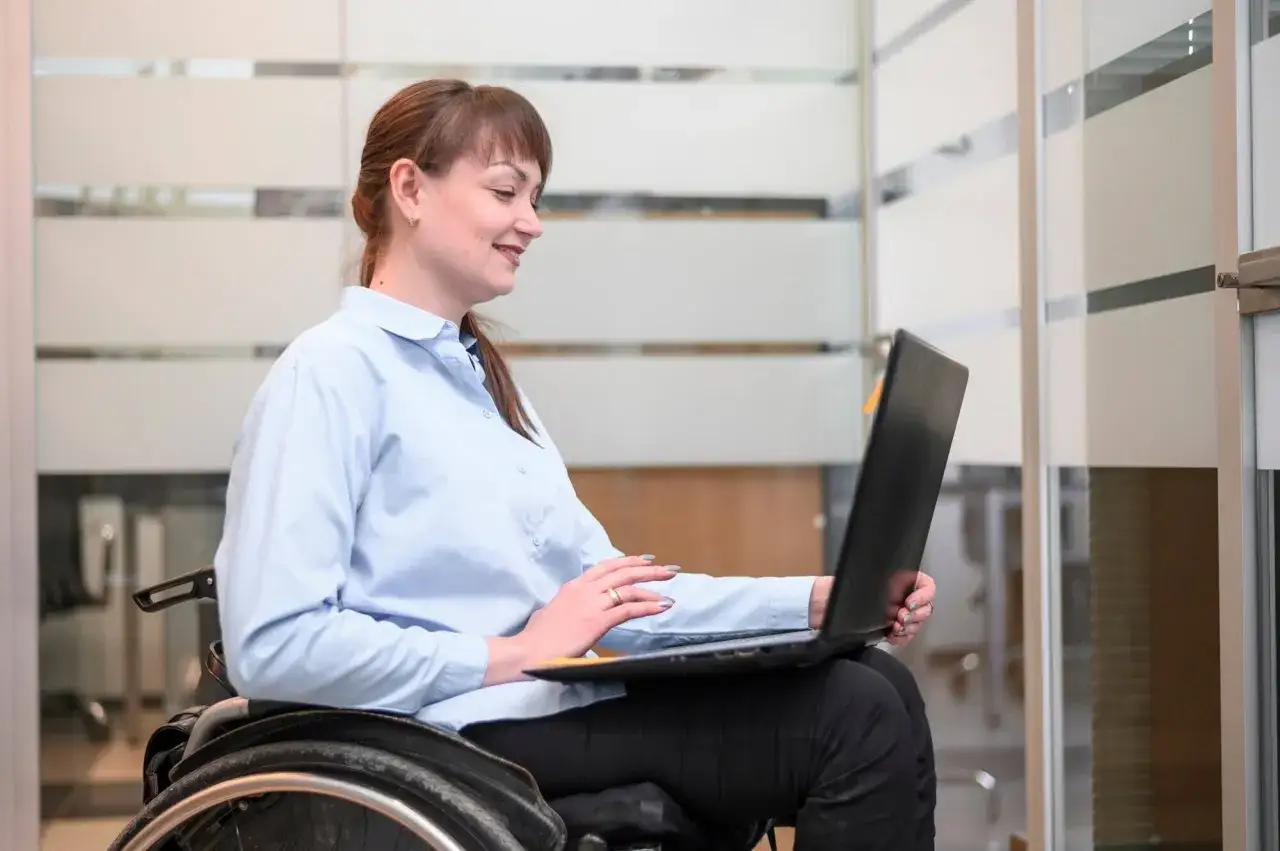 Femme souriante en fauteuil roulant, rqth déjà en poste, travaillant sur son ordinateur portable dans un bureau moderne.