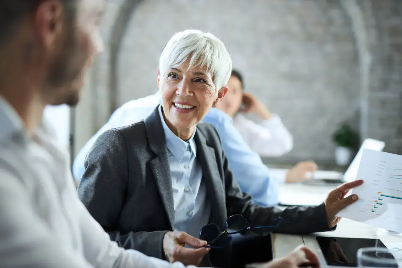 Une femme souriante discute d'un graphique avec un coll&egrave;gue, peut-&ecirc;tre pour pr&eacute;parer sa mise &agrave; la retraite par l'employeur.
