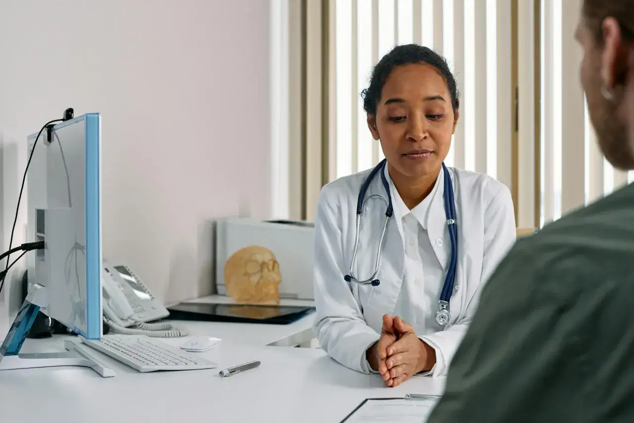 Médecin écoutant attentivement un patient lors d'une visite de reprise obligatoire. Bureau médical avec ordinateur, téléphone et un crâne en décoration.